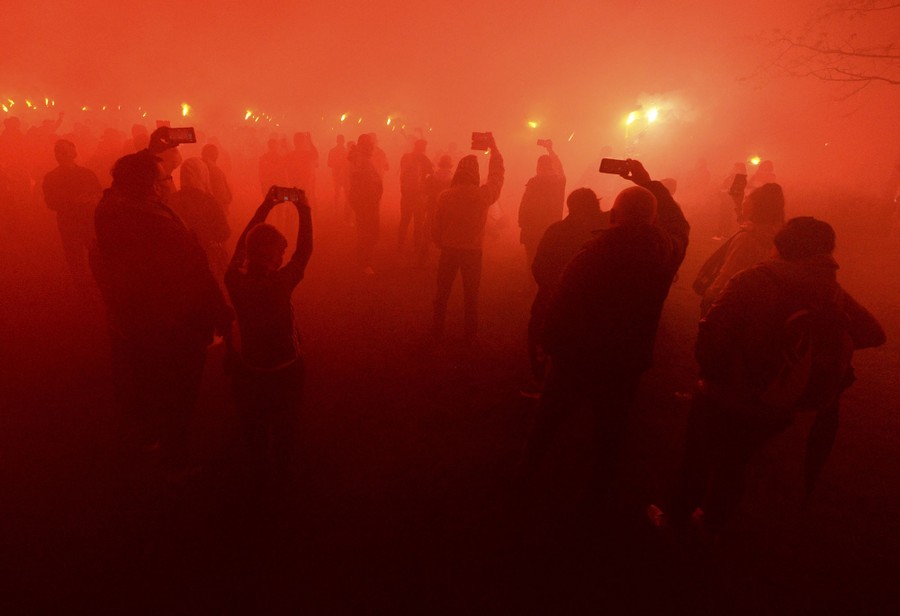 Dozens of people stand in a smoky haze, holding up their phones and flares.