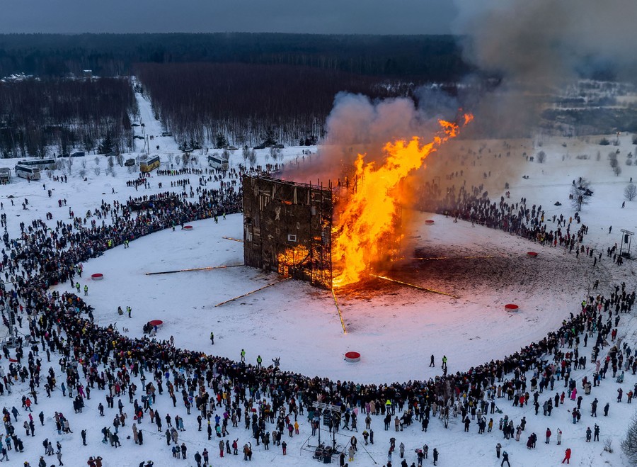 A large crowd stands in a circle around a tall, burning, cube-shaped installation.