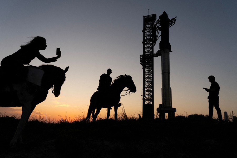 Two people on horseback take photos near a tall rocket on a launch pad.
