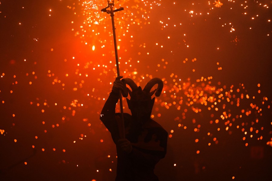 A person wearing a horned mask carries a staff in a dark street with many fiery sparks in the air beyond them.
