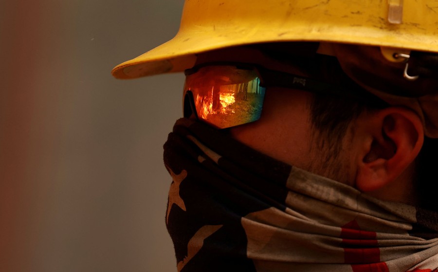 A firefighter wearing a mask and sunglasses looks toward a forest fire.