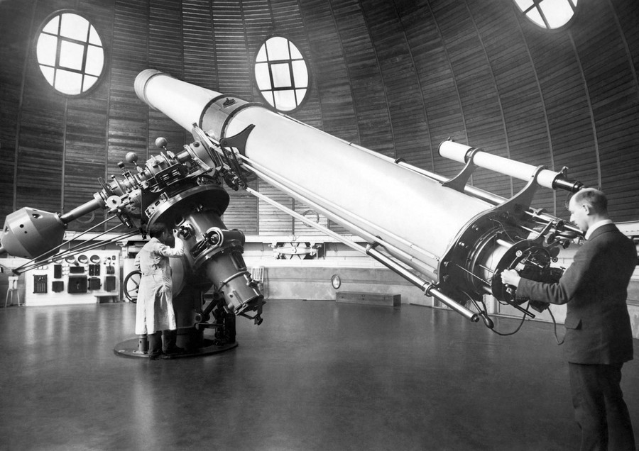 Two people operate a very large telescope inside a domed observatory.