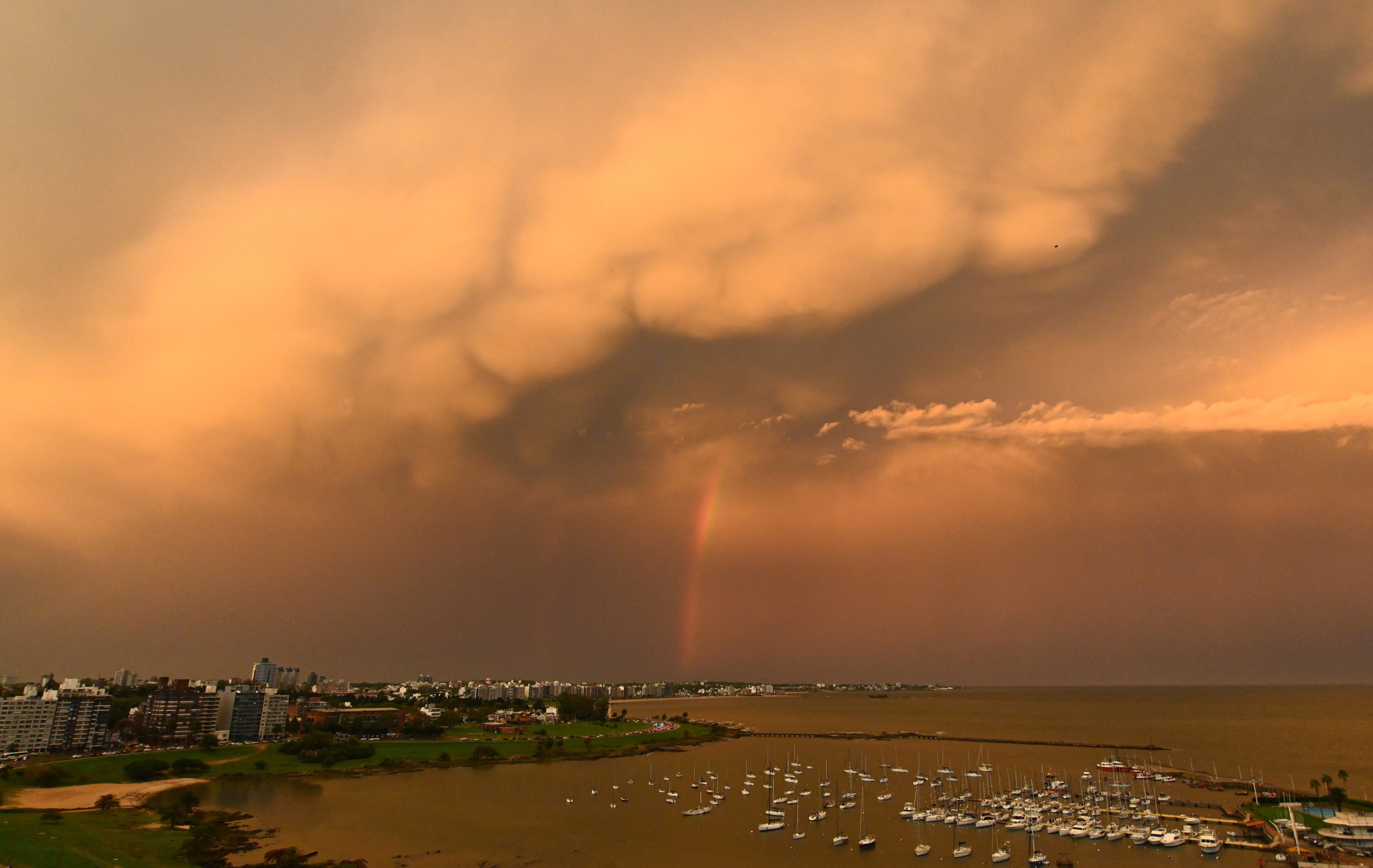 A rainbow appears beneath clouds lit by the setting sun over a harbor.