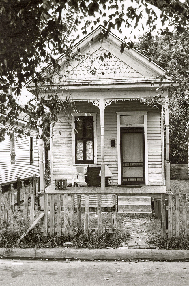 black-and-white photo of small wood-sided house with front porch framed by trees