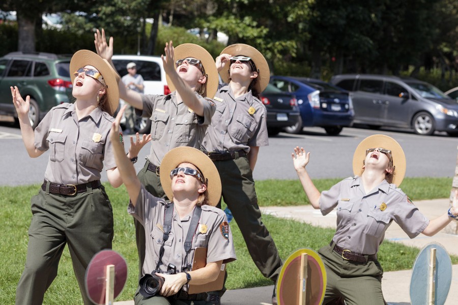 A group of five park rangers wearing eclipse glasses look skyward and raise their arms playfully.