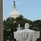 Photograph of a statue from the backside holding a sword. The U.S. Capitol Building is in the background, shrouded by trees.