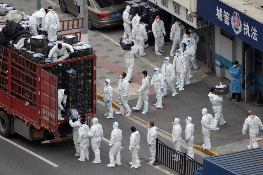 Two lines of people wearing protective gear stand on a sidewalk and street, unloading food supplies from a truck.