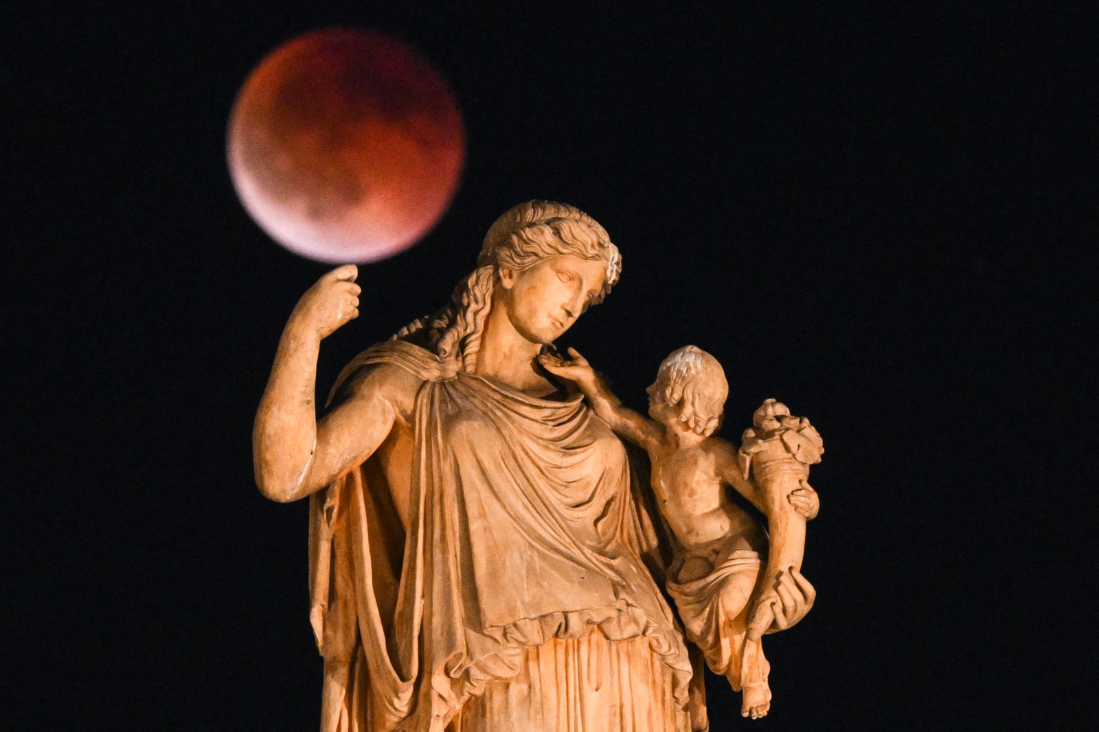 A red-colored full moon is seen during an eclipse behind a statue.