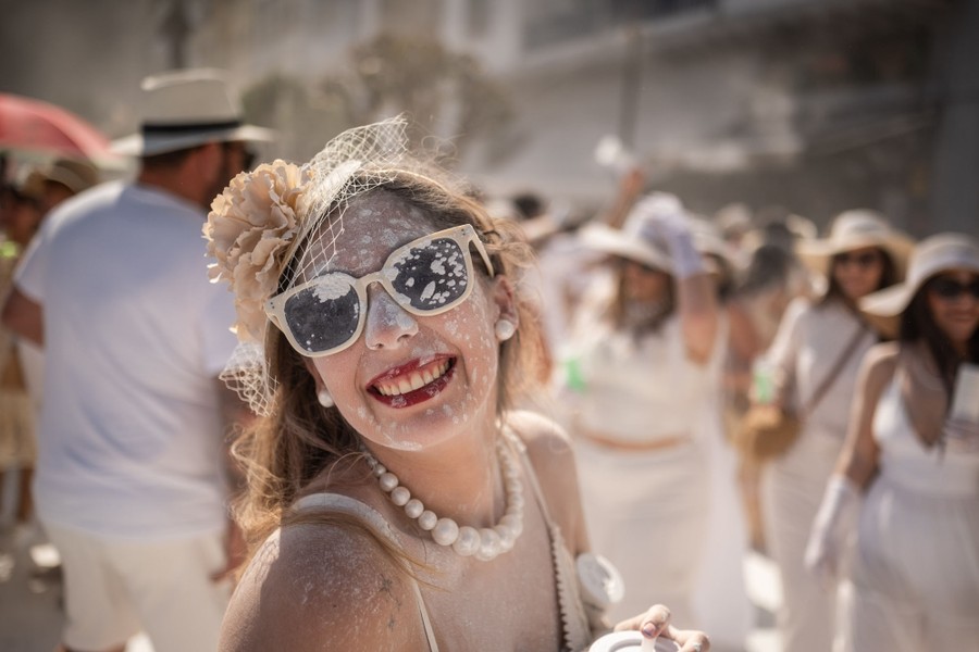 A person smiles, among others, all wearing white, and partly-covered in thrown talcum powder.