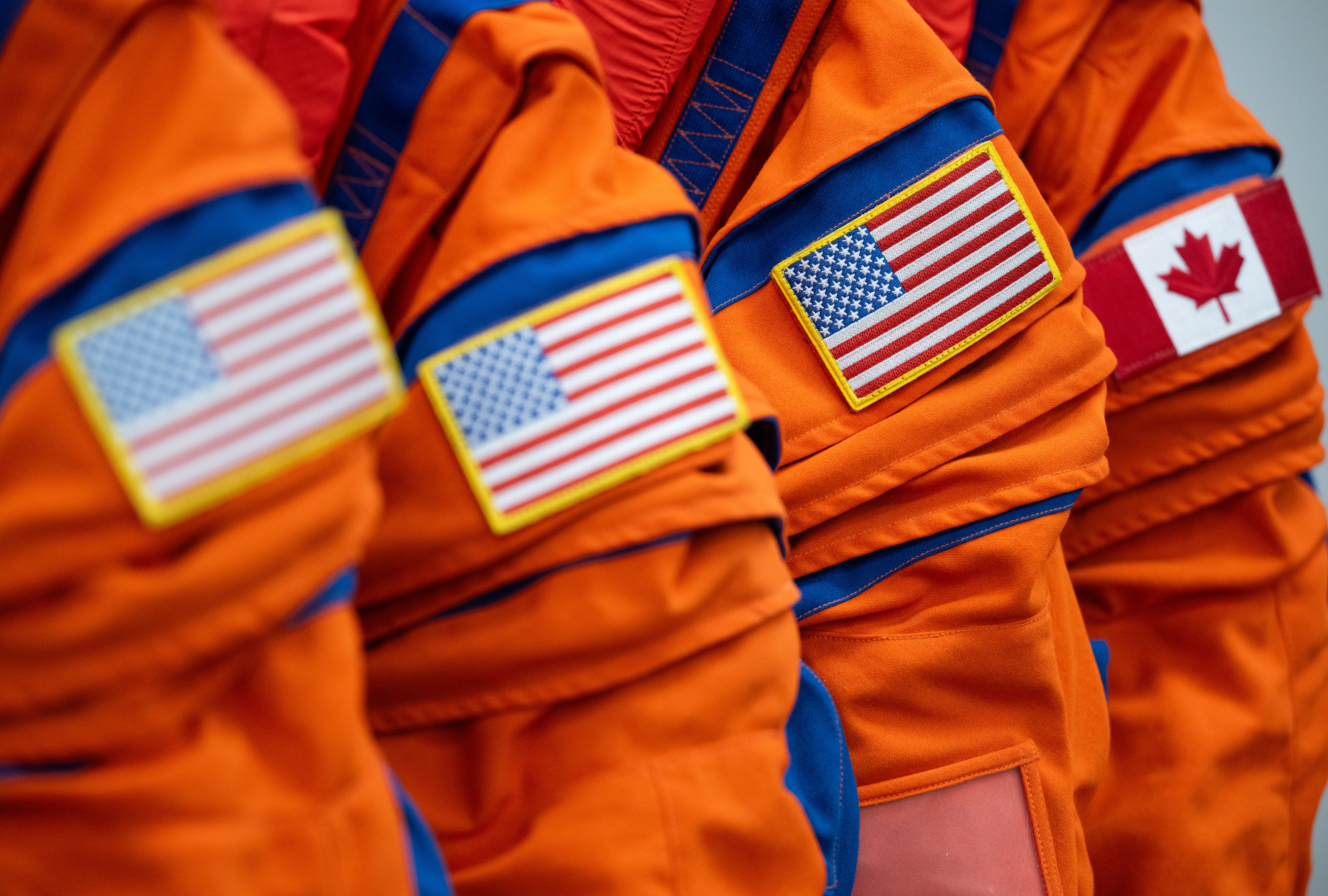 Three American flag arm patches and one Canadian flag arm patch are seen on the shoulders of four orange flight suits.