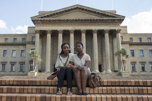 Two women sit with linked arms on brick steps outside a stately building with columns.