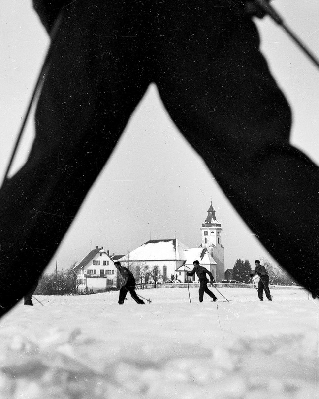 A black and white photograph of a winter scene of skiers, seen through the outstretched legs of another skier