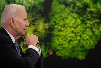 President Biden, in profile in front of a wall of green plants, gazes forward with his hands clasped.