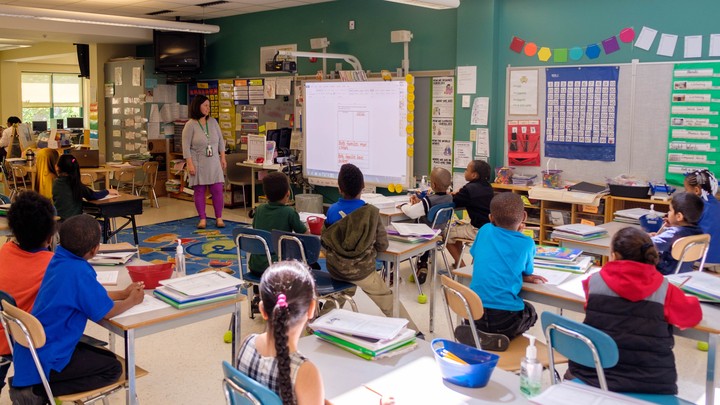 First-grade students are seated in a classroom listening to their teacher.