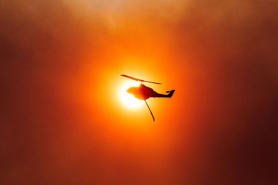 A firefighting helicopter is silhouetted in front of the sun in a smoky sky.