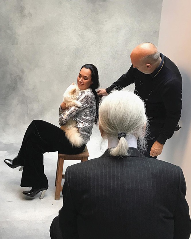 photo of photo shoot with gray backdrop and woman sitting on chair with fluffy cat in her arms, one man helping to pose her, while a an in black jacket and white ponytail is looking on seen from the back
