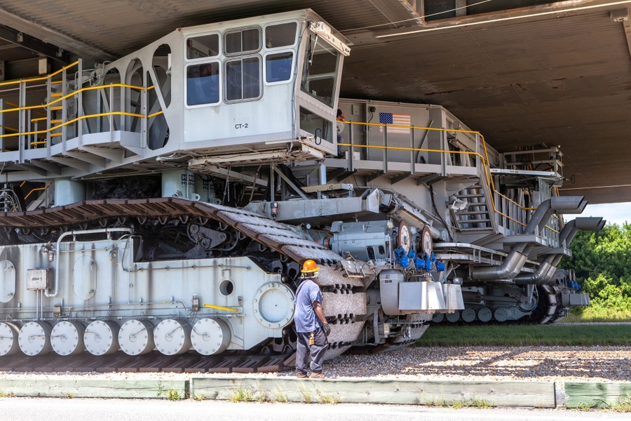 A person stands beside the tracks of the massive crawler-transporter as it slowly carries a rocket.