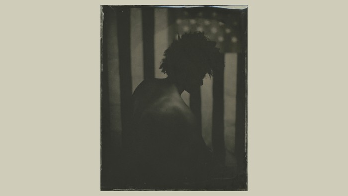 A photograph of a Black man, his back facing the camera, with a large American flag behind him