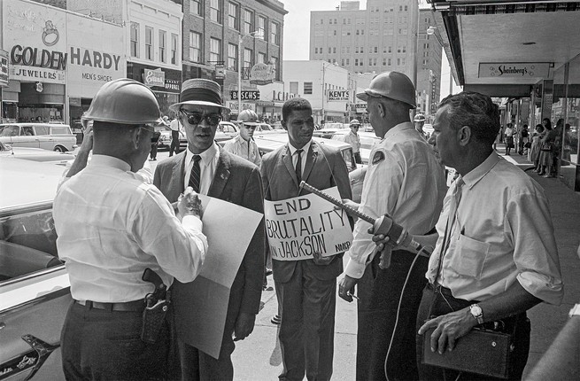 black and white photo of two Black men in suits, ties, and fedoras on a downtown street, one of them holding a sign saying "END BRUTALITY IN JACKSON," surrounded by 3 white men, two in hard hats and police gear