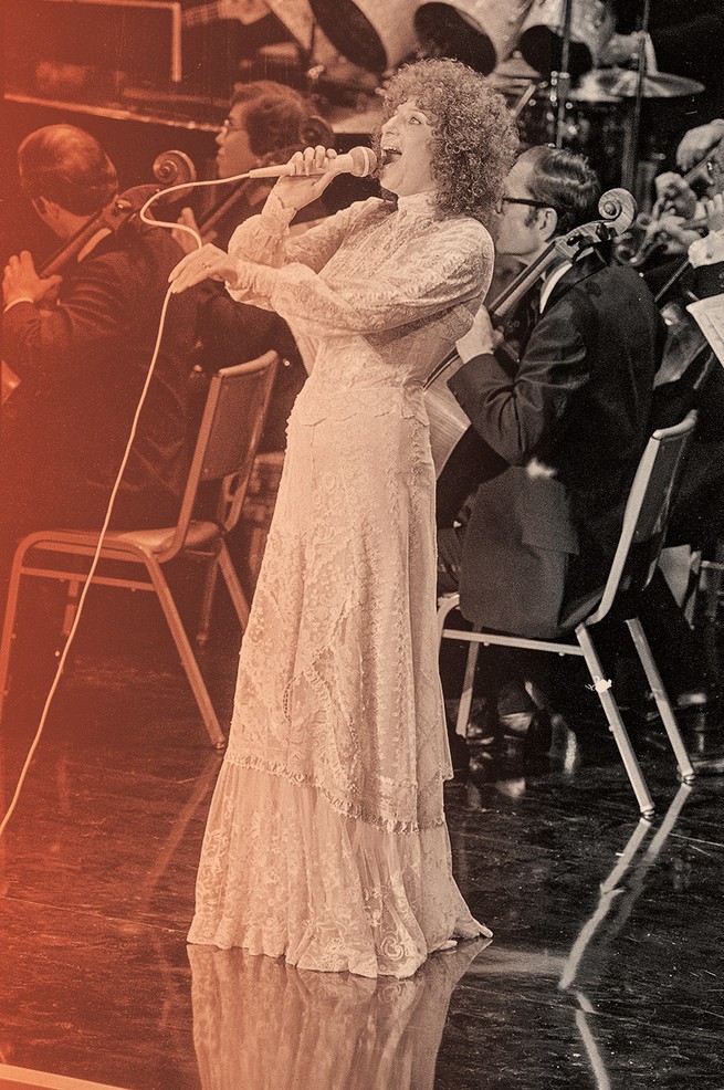 photo of Barbra Streisand in white gown on stage singing into microphone with orchestra in background