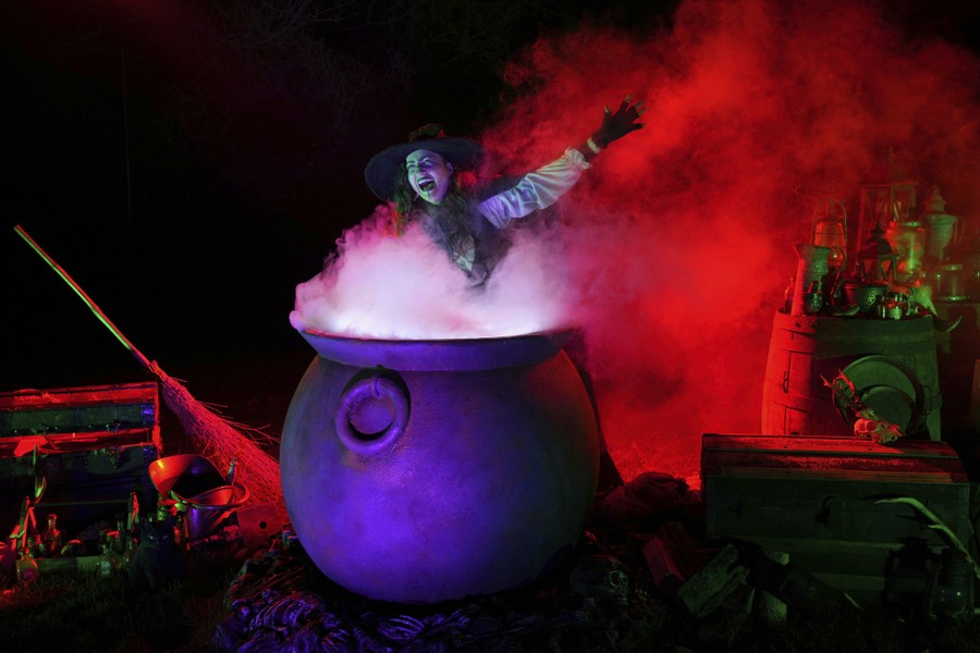 A model dressed as a witch poses behind a "smoking" cauldron prop.