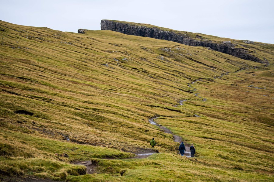 Several people hike on a mountainside trail, near sea cliffs.