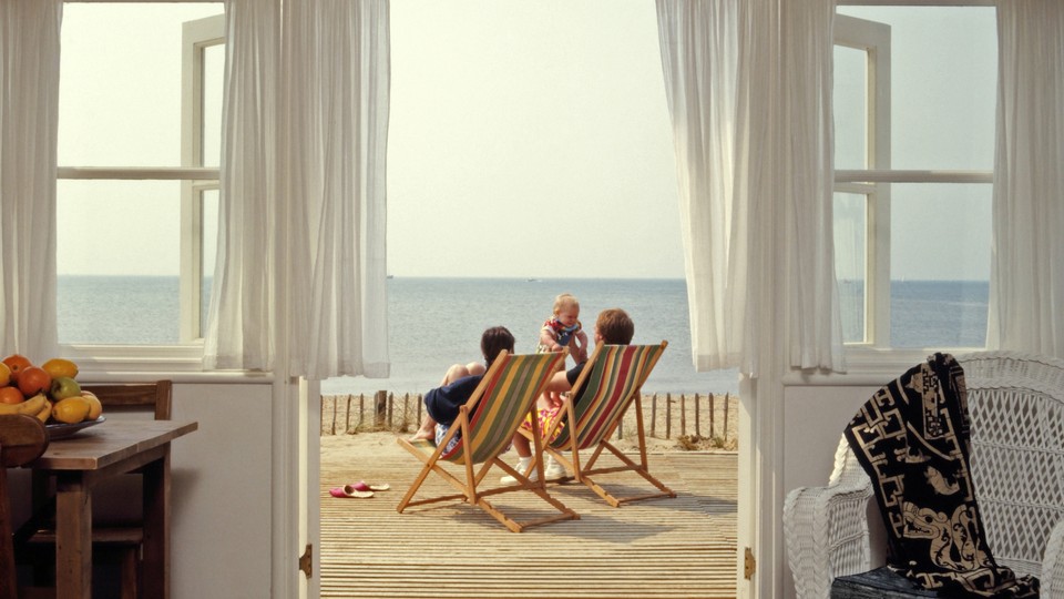 A hazy photograph of two people in beach chairs looking onto the ocean, taken through open patio doors from inside a house