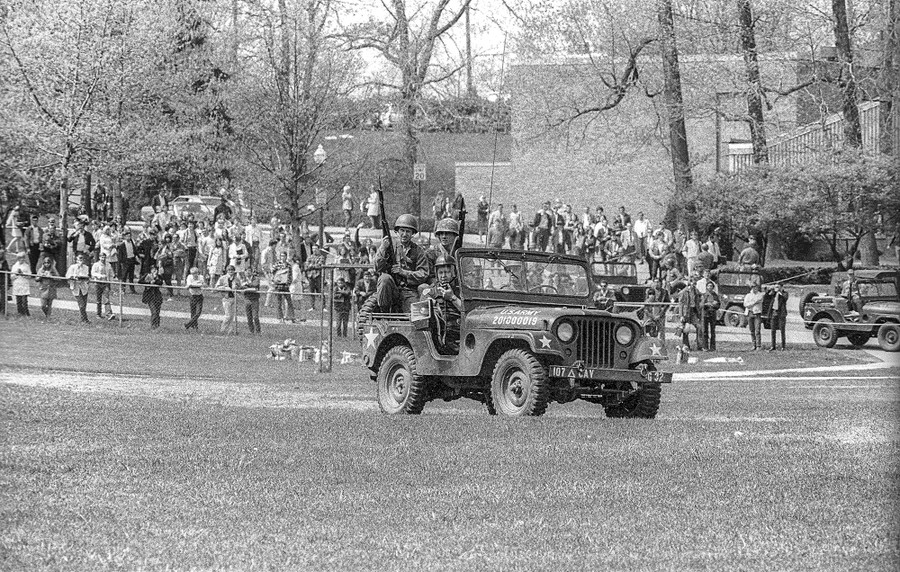 A police officer and three National Guardsmen drive a jeep on a lawn, with many onlookers in the distance.