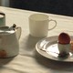 A table covered in a white table cloth, and on top, a tea kettle, two mugs, and a plate with an egg, a spoon, and a slice of bread