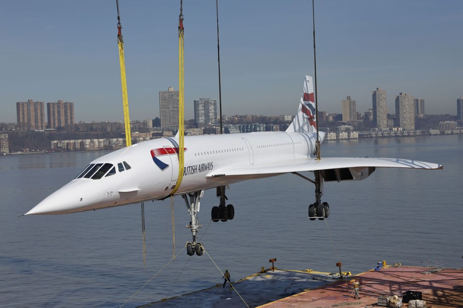 A large aircraft is lifted off a barge by a crane using long straps.