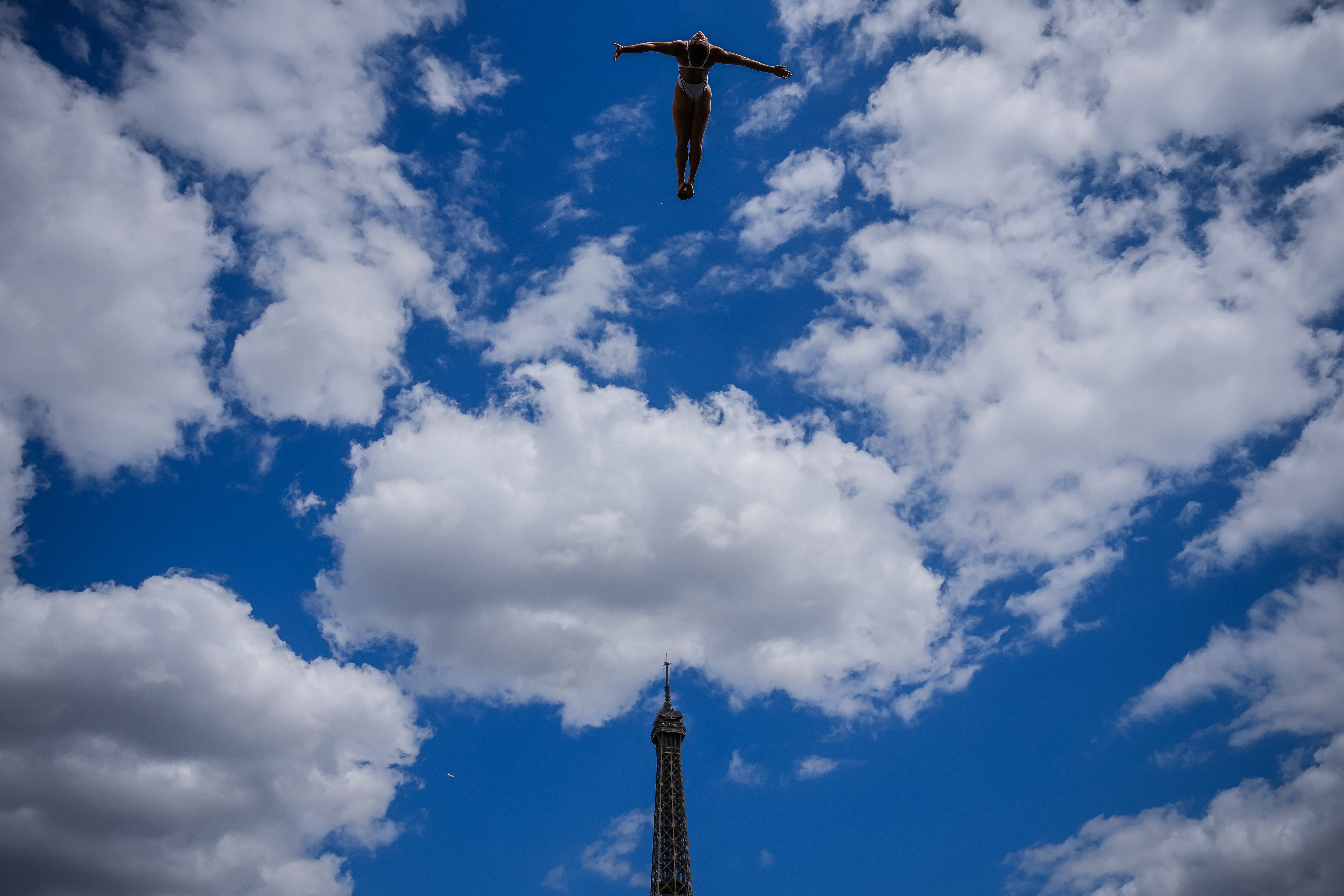 A high diver falls under a cloud-filled sky, with the Eiffel Tower seen in the background.