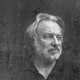 Black and white photo of a man in a black short sleeved shirt with a gray beard and swooping hair.