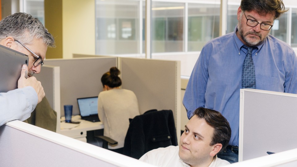 Three male employees of a news website sit and stand in cublcles.