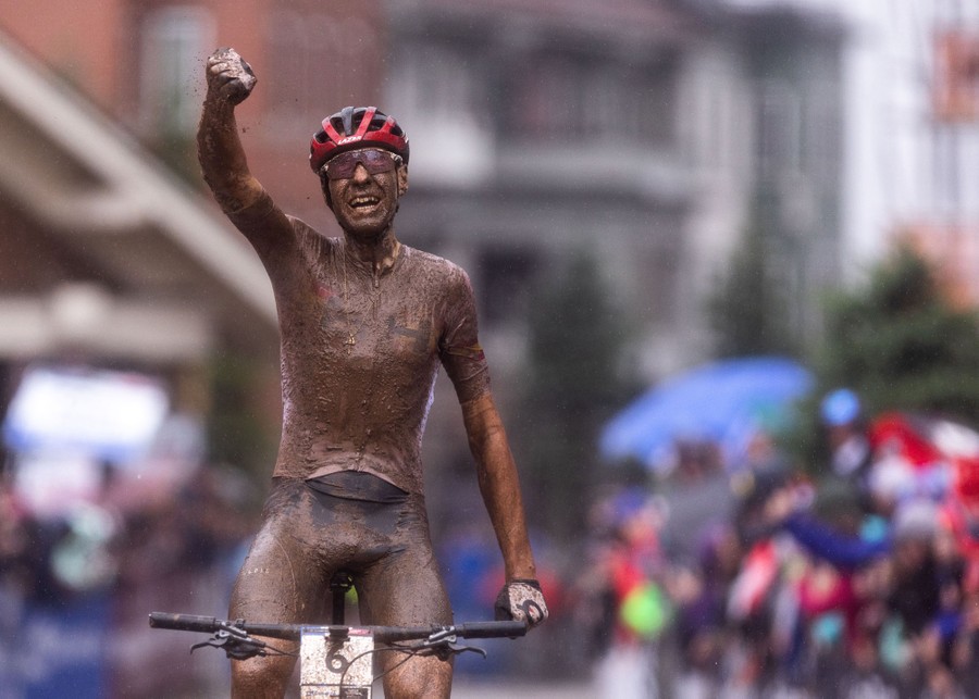A mud-covered cyclist raises a fist while crossing a finish line.