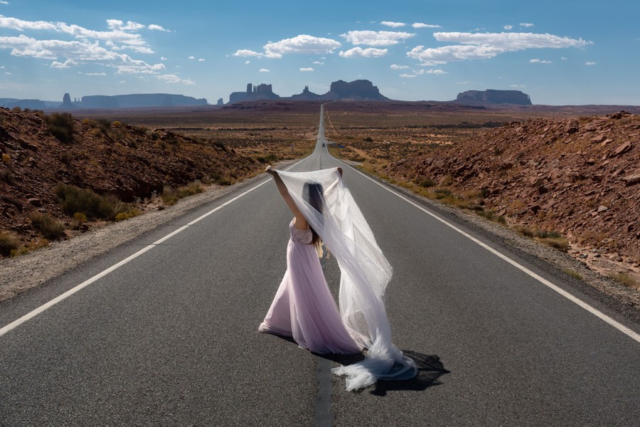 A person in a wedding dress poses for a photo, standing in the middle of a two-lane road in the desert, with picturesque buttes rising in the distance.
