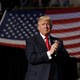 President Donald Trump stands in front of an American flag with clasped hands.