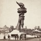 People gather at an information booth at the base of a very tall statue of an arm holding a torch, in a photo from 1876.