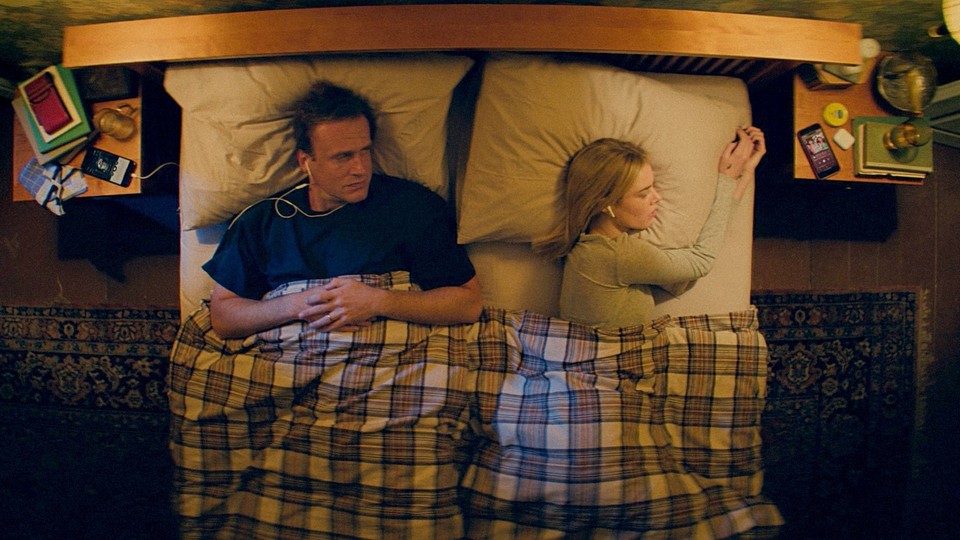 Jason Segel looks at Samara Weaving while lying in a bed with a plaid comforter