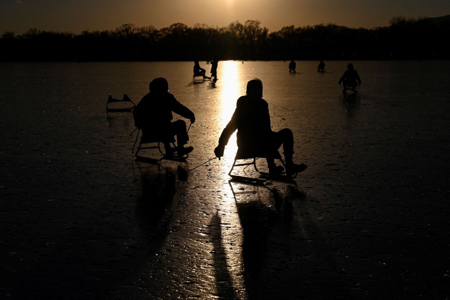 Several people slide on a frozen lake, using sticks to push themselves along while sitting in chairs on ice blades.
