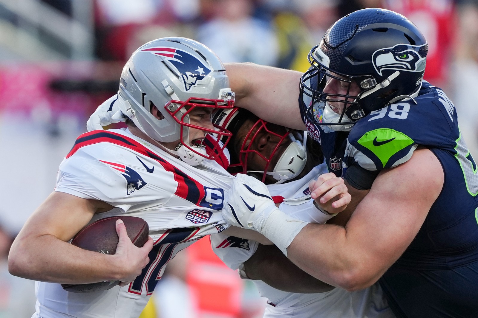 A Seahawk defensive football player grabs the Patriots quarterback to sack him, crushing a Patriots offensive lineman in between them.
