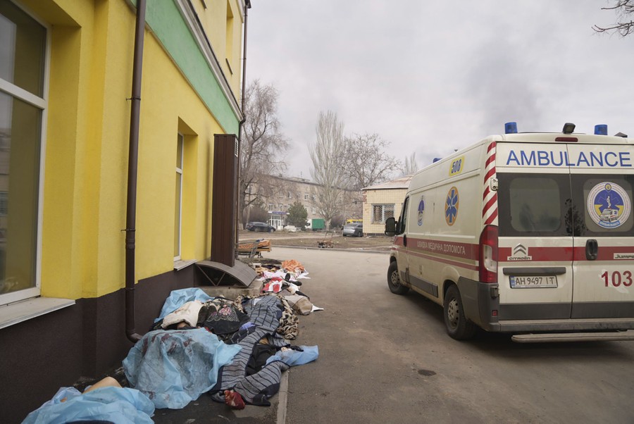 An ambulance is parked near a building, with an unknown number of bodies (perhaps a dozen) covered by blankets and sheets, on the ground beside the building.