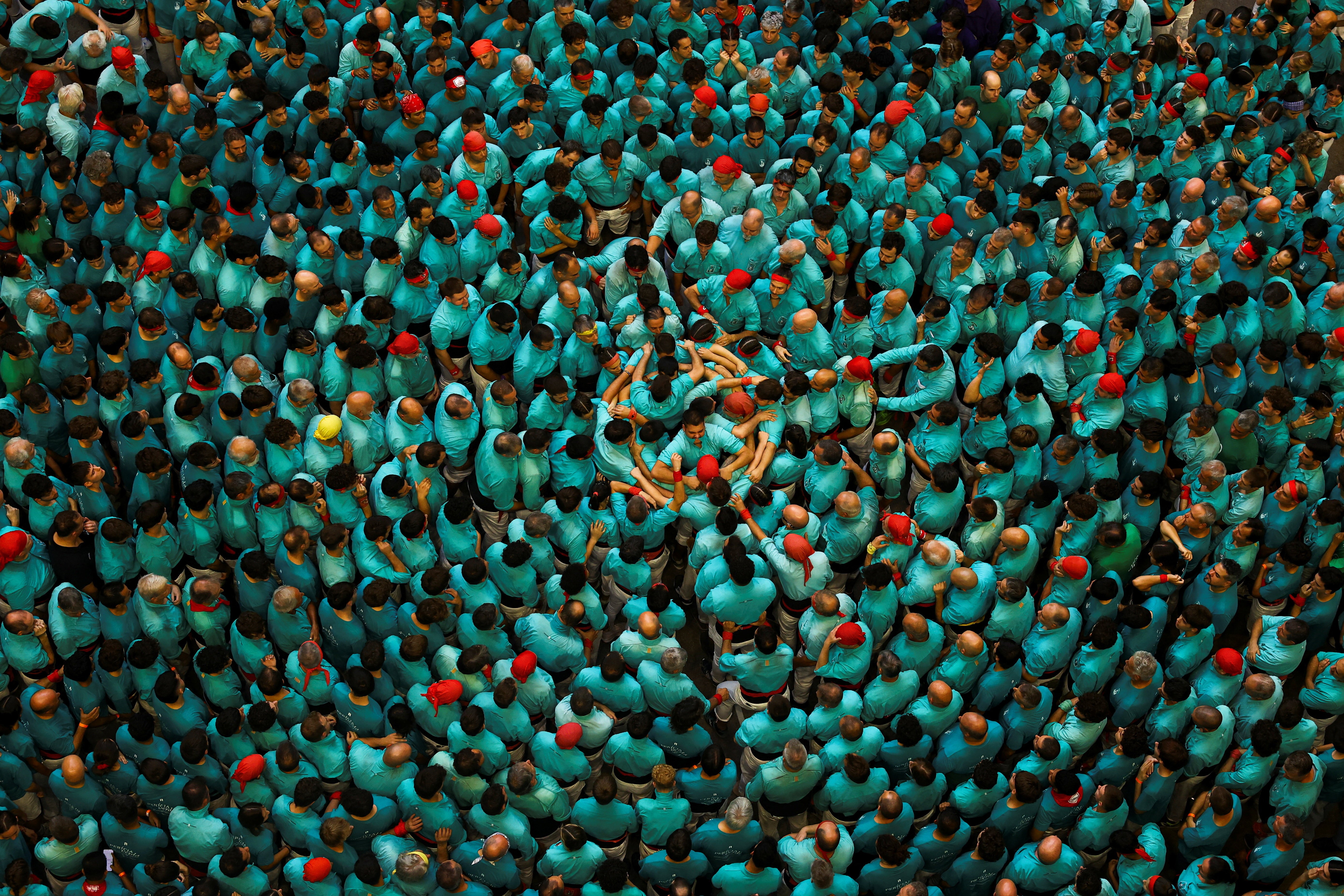 An overhead view of a large crowd of people wearing blue-green shirts, gathered in roughly concentric circles, smashing together, preparing to build a human tower