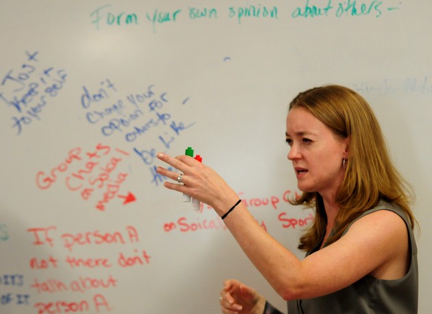 A woman stands at a dry-erase board with phrases written on it. She has markers in her hand.