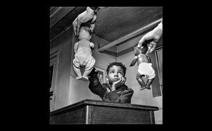 Black and white photo of two hands offering dolls to child seated at desk, child reaching for the white doll while looking at the Black doll