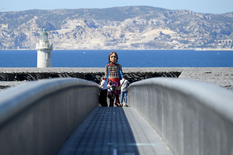 A giant puppet walks across a pedestrian bridge, with ocean and mountains visible in the background.