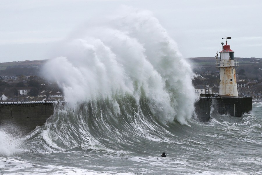 A person swims and takes photographs near large waves crashing against a pier.