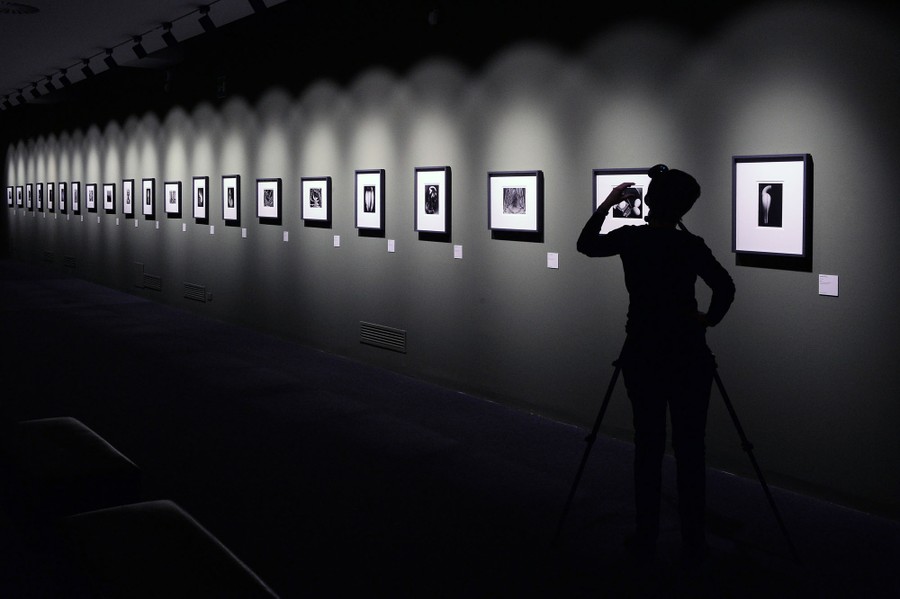 A person stands in a gallery, viewing a couple dozen framed photographs hung along a long wall.