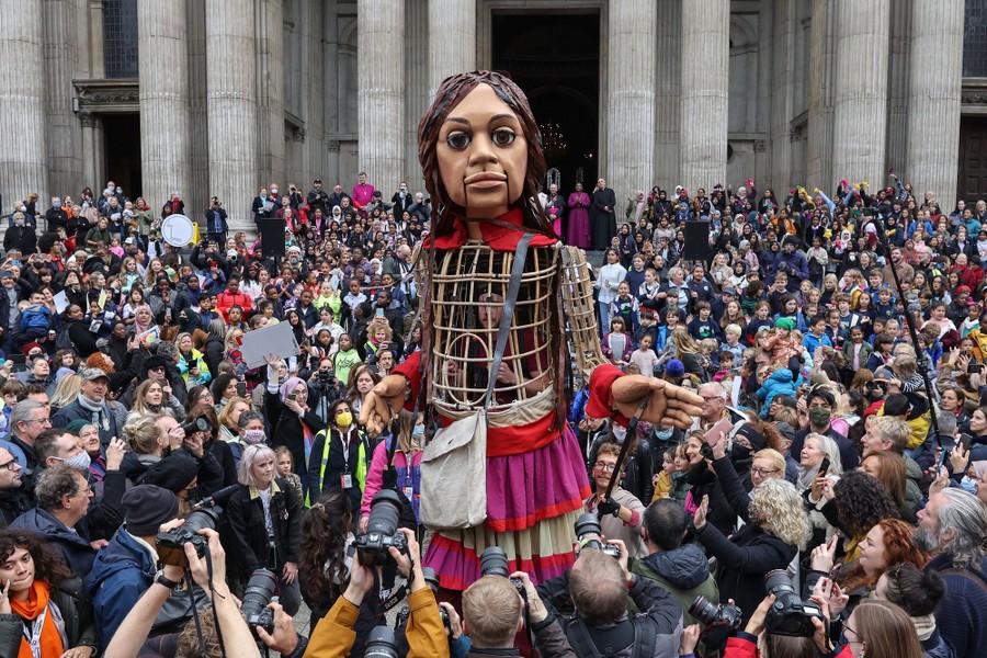 A tall puppet depicting a young girl stands among a large crowd outside a cathedral.