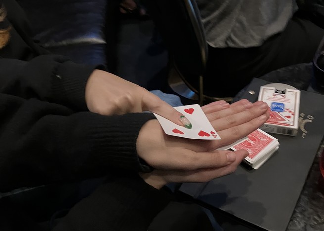 A deck of playing cards, two sets of hands. One person is holding a playing card—the four of hearts—on top of the other person's hand, face up.