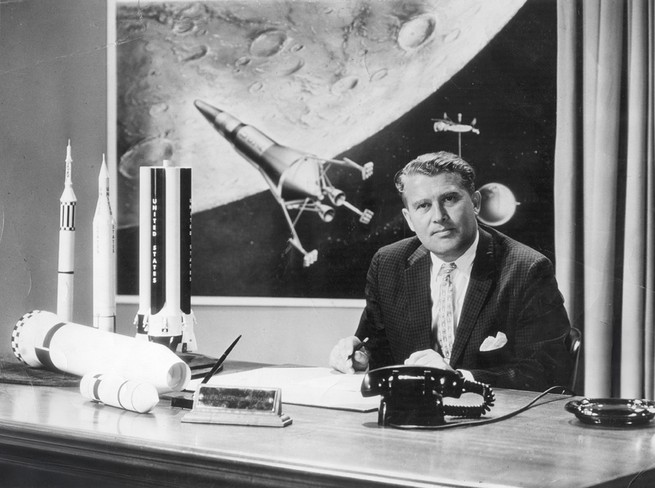 black-and-white photo of man in suit and tie sitting behind desk, with rotary telephone, ash tray, and several rocket models, with a poster-size drawing of a rocket near the moon behind